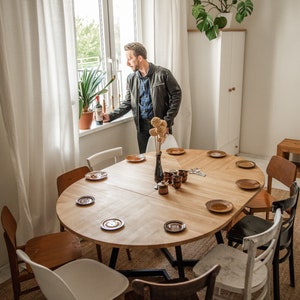 May include: A round wooden dining table with eight chairs, set with plates and cups. A man stands by a window holding a bottle of wine.
