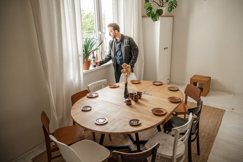 May include: A round wooden dining table with eight chairs, set with plates and cups. A man stands by a window holding a bottle of wine.