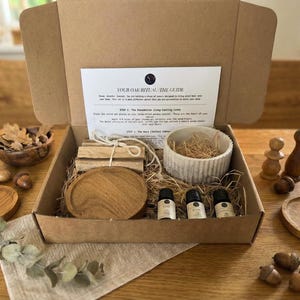 Może przedstawiać: Otwarty karton zawierający drewniany zestaw rytualny. Zestaw zawiera drewniane przedmioty, butelki z olejkami eterycznymi i przewodnik. Dodatkowe drewniane przedmioty i żołędzie są ułożone wokół pudełka na drewnianym stole. Tekst na przewodniku brzmi "YOUR OAK RITUAL: THE GUIDE".