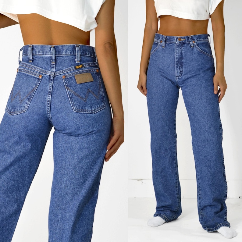 May include: A pair of high-waisted, straight-leg jeans in a classic blue denim wash. The jeans have a relaxed fit and feature a brown leather patch with the Wrangler logo on the back.