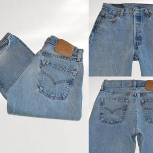 Könnte beinhalten: Hellblaue Jeans aus Denim mit Knopfleiste, Fünf-Taschen-Stil und einem beigen Lederaufnäher am hinteren Bund. Das Design umfasst kupferfarbene Nieten, ein kleines rotes Etikett an der Gesäßtasche und klassische bogenförmige Nähte.