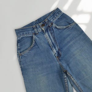Puede incluir: Vaqueros de mezclilla azul claro con un diseño clásico de cinco bolsillos. Los vaqueros tienen cierre de botón y cremallera, trabillas para el cinturón y un lavado descolorido para un look vintage. Las costuras son visibles.