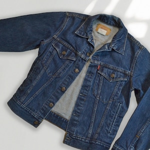 May include: A classic blue denim jacket with a button-up front, two chest pockets, and a collar. The jacket features copper-colored buttons and a red tag on the chest pocket. The jacket is made of a sturdy denim material.