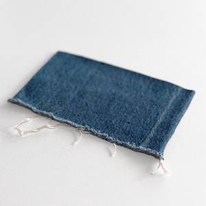 May include: A rectangular piece of denim fabric in a medium blue hue. The fabric has a raw, frayed edge with loose white threads. The texture of the denim is visible, suggesting a worn or distressed appearance. The item is likely a patch or a piece of fabric for crafting.