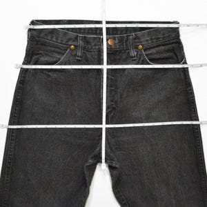 May include: Black denim jeans with a straight leg, featuring a button and zipper closure, and classic five-pocket styling. A measuring tape is laid across the jeans to show the waist and length measurements.