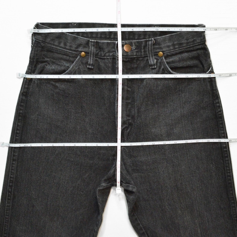 May include: Black denim jeans with a straight leg, featuring a button and zipper closure, and classic five-pocket styling. A measuring tape is laid across the jeans to show the waist and length measurements.