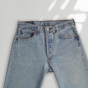 May include: Light blue denim jeans featuring a classic five-pocket design, a button fly closure with a metallic button, and copper-toned rivets at the pocket corners. The fabric displays a faded wash with subtle whiskering and light distressing.