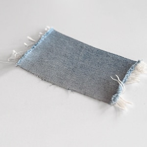 May include: A rectangular swatch of denim fabric with frayed edges. The denim is a medium blue color with a diagonal weave pattern. The edges are unfinished, with white threads extending from the fabric.