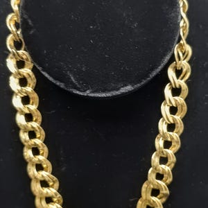 May include: A gold-colored chain necklace with a thick, interlocking link design. The necklace is displayed on a black velvet surface, showcasing its shiny, metallic appearance. The chain is a statement piece, suitable for various occasions.