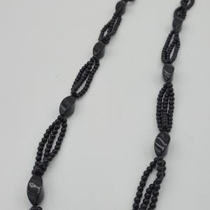 May include: A black beaded necklace with three strands of small, round beads. The necklace features larger, oval-shaped beads spaced along the strands. The necklace is displayed against a white background.