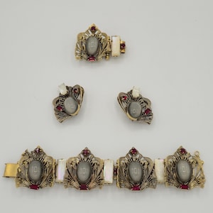 May include: A vintage jewelry set featuring a bracelet, a pair of earrings, and a clasp. The set has a gold-tone metal with gray oval center stones, red accents, and white rectangular stones. The bracelet has multiple decorative panels.