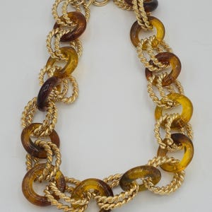 May include: A gold-tone necklace featuring a series of amber-colored, circular links. The links are connected by twisted gold-tone metal. The necklace has a toggle clasp closure. The necklace is a statement piece.