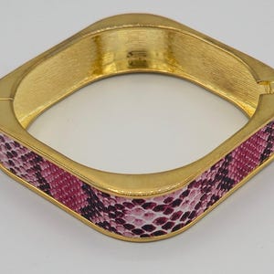 May include: A gold-toned square bangle bracelet with a pink and purple snakeskin pattern. The bracelet has a hinged clasp and a brushed gold finish. The snakeskin pattern wraps around the bracelet.