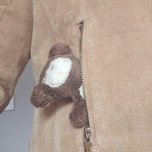 May include: A light tan suede jacket featuring a vertical zipped pocket. A small, brown and white plush bear toy is tucked inside the pocket, with its head and upper body visible, showing a textured fabric surface and a metallic zip pull.