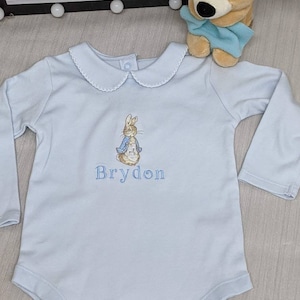 May include: A light blue baby onesie with a Peter Rabbit embroidery and the name "Brydon" embroidered below.
