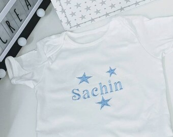 May include: A white short-sleeved baby bodysuit featuring the embroidered name Sachin in light blue thread, flanked by two light blue embroidered stars. The garment is positioned below a white storage bin decorated with a grey star pattern.