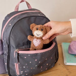 May include: A dark grey backpack with pink trim, featuring a star and heart pattern, a side mesh pocket with a light purple water bottle, and a small brown plush teddy bear with a pink heart patch being placed into the front zippered pocket.