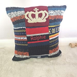May include: A decorative cushion with a crown design and the name "Ralphie" embroidered in red. The patchwork design incorporates various knit patterns in blue, orange, white, and black, offering a textured, cosy feel.