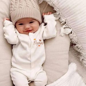 May include: A white baby onesie with long sleeves and a button fastening. The onesie has a small embroidered design of leaves and the name "Flynn" on the front. The baby is wearing a beige knitted hat.