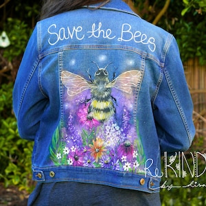 Könnte beinhalten: Blaue Jeansjacke mit handgemaltem Design mit einer großen Biene, Blumen und dem Schriftzug "Save the Bees" in weiß. Die Jacke hat eine Knopfleiste und einen klassischen Kragen. Das Kunstwerk ist lebendig und detailliert.