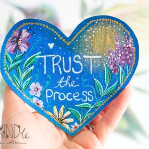 Op de afbeelding: Een handbeschilderde hartvormige patch met een blauwe achtergrond en de woorden "Trust the Process" in het wit. De patch heeft bloemen- en bladmotieven in paars, groen en geel, met een gouden rand. De patch wordt in een hand gehouden.