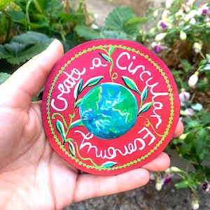 Op de afbeelding: Een ronde houten schijf, rood geschilderd met een centrale illustratie van de aarde in blauw en groen. De tekst "Create a Circular Economy" staat in witte letters rond de wereldbol, geaccentueerd door groene ranken en een gestippelde groene rand.