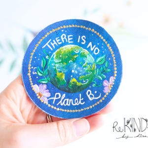 May include: A circular blue patch with the text "THERE IS NO Planet B". The patch depicts a painted Earth, surrounded by green leaves and purple flowers, with a gold beaded border.