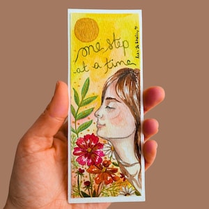 May include: A watercolour bookmark with a yellow background and the text "one step at a time." The bookmark features an illustration of a woman smelling flowers. The flowers are red, orange, and white.