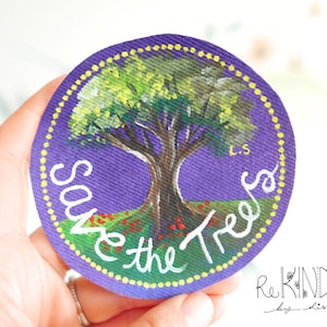 May include: Round, purple patch with a hand-painted tree design and the words "Save the Trees" in white. The tree has green leaves, a brown trunk, and red flowers at its base. The patch is edged with yellow dots.