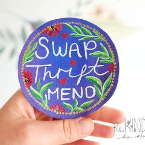 May include: A round, blue patch with the words "SWAP, THRIFT, MEND" in white script. The patch is decorated with red flowers, green leaves, and a gold border. The patch is held in a hand.
