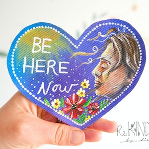 May include: Heart-shaped artwork with the words "BE HERE Now" in white lettering. The artwork features a profile of a person with flowing hair, flowers, and a blue and yellow gradient background. The artwork is held by a hand.