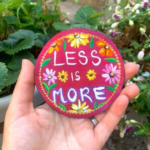 May include: A circular wooden plaque painted in a deep red hue, featuring the white hand-painted text "LESS IS MORE" at the centre. The design is surrounded by a decorative border of colourful painted flowers in yellow, orange, and pink tones.