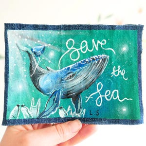 Op de afbeelding: Een handgeschilderde patch met een blauwe walvis met de woorden "Save the Sea" in witte letters. De walvis staat tegen een teal en groene achtergrond met witte accenten, omlijst door een donkerblauwe rand.