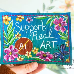 May include: A rectangular fabric patch with a teal background featuring hand-painted flowers, green leaves, and a blue butterfly. The text reads Support Real ART, with a brown circle containing a diagonal line over the letters AI.