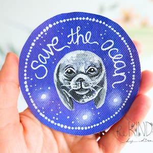 Puede incluir: Un parche redondo azul con un borde punteado blanco y las palabras "Save the ocean" en escritura blanca. El parche presenta una ilustración de foca pintada a mano en el centro.