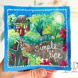 May include: Square fabric art with a blue border and a painted landscape scene. The artwork features a small house, trees, flowers, and the words "simple life". The painting is on a blue fabric with a white dotted border.