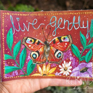 Op de afbeelding: Een handgeschilderd kunstwerk op een bordeauxrode achtergrond toont een vlinder met rode, oranje en zwarte vleugels. De woorden "live gently" zijn geschreven in lichtblauw script. Groene bladeren en kleurrijke bloemen omringen de vlinder.