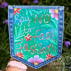 May include: Hand-painted denim pocket with the message "Say No to Fast Fashion" in colorful script. The pocket is decorated with flowers and leaves in shades of red, pink, and green against a teal and green background. The pocket is framed with blue denim.