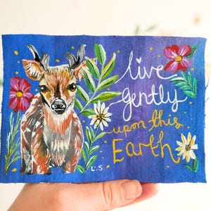 Może przedstawiać: Ręcznie malowana niebieska naszywka z tkaniny przedstawiająca jelenia z rogami, otoczonego kwiatami i liśćmi. Tekst "live gently upon this Earth" jest napisany białą i żółtą czcionką. Grafika jest podpisana "L.S."