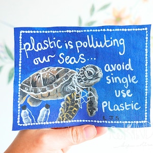 Op de afbeelding: Een blauw canvas met een witte stippellijnrand toont een geschilderde zeeschildpad en de tekst "plastic is polluting our seas... avoid single use plastic." De schildpad is bruin, zwart en wit.
