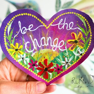 Pode incluir: Uma obra de arte em forma de coração com as palavras "be the change" em escrita branca. O fundo roxo é decorado com flores amarelas, vermelhas e brancas e folhagem verde. A obra de arte é segurada por uma mão.