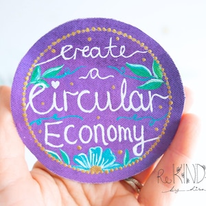 May include: A round, purple fabric patch with the words "create a circular economy" in white script. The patch is decorated with teal and green leaves, teal accents, and a gold dotted border. The patch is held in a hand.