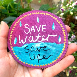 May include: A circular fabric patch featuring a purple background with a gold dotted border. The text "SAVE water" is written in white, and "save life" is written in black over a blue wave graphic. Four light blue water droplets are depicted.