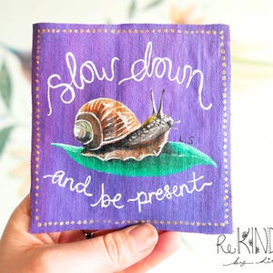 Könnte beinhalten: Ein lila Quadrat mit dem weißen Schriftzug "slow down and be present". Auf dem Quadrat sind eine Schnecke mit braunem Gehäuse und ein grünes Blatt gemalt. Die Ränder sind mit goldenen Punkten versehen.
