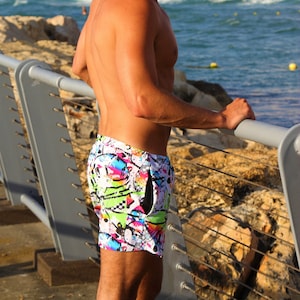 May include: A man wearing colorful swim trunks with a white background and a variety of graphic designs. He is standing on a wooden pier with a metal railing.
