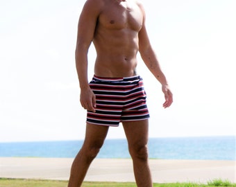 May include: A man wearing white sneakers and red, white, and blue striped swim trunks. He is standing on a grassy surface with the ocean in the background.