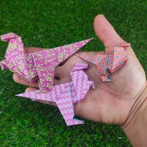 May include: Three origami dinosaurs made of paper. One is pink with a green floral pattern, one is pink with a white chevron pattern, and one is peach with a blue floral pattern.