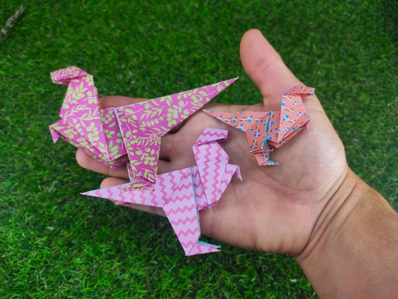 May include: Three origami dinosaurs made of paper. One is pink with a green floral pattern, one is pink with a white chevron pattern, and one is peach with a blue floral pattern.