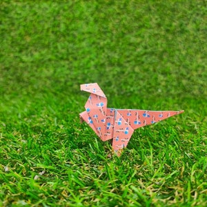 May include: A pink origami dinosaur with a blue and white floral pattern stands on a green grassy surface.
