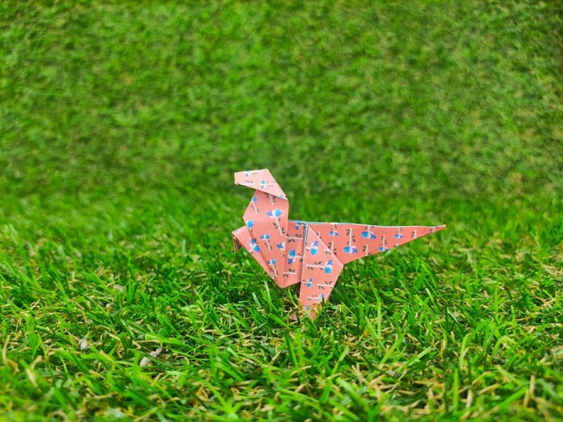 May include: A pink origami dinosaur with a blue and white floral pattern stands on a green grassy surface.